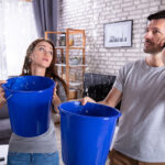 Couple Using Bucket For Collecting Water Leakage From Ceiling And Calling Plumber On Cellphone After Having Issues With The Plumbing In Their New Home